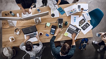 An overhead shot of a group working at a messy table full of computers and papers.