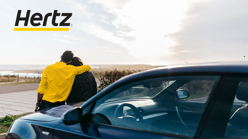 Hertz logo superimposed on scene of couple with rental car.
